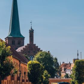 Blå himmel over en charmerende, orange købstad, med grønne træer og et kirketårn med grønt spir i baggrunden