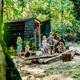 Fem børn og to voksne samlet om et bål i skoven. Bag ved dem ses et arkitekttegnet shelter.