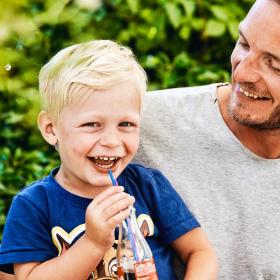 En lille lyshåret dreng sidder på skødet af sin far. Drengen har en blå t-shirt på og en cola med sugerør i hånden. Han smiler stort til kameraet, men hans far kigger smilende ned på ham. De er omgivet er en grøn hæk.