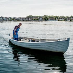 Hvid båd med mand i ternet skjorte ombord sejler ud fra Kerteminde Havn på roligt vand. Byen ses i baggrunden i horisonten.