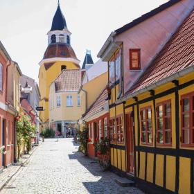 En malerisk gade i Faaborg med brostensbelægning og farverige huse med bindingsværk. Husene har levende gule og røde facader med traditionelle røde tegltage. I baggrunden tårner Faaborgs karakteristiske klokketårn sig op med sin røde kuppel og spir, hvilket tilføjer et charmerende historisk element til scenen. Det er en solrig dag med blå himmel og stokroser i fuldt flor, der tilføjer liv til gadebilledet.