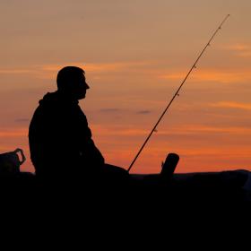 Sorte silhuetter af to skikkelser, der sidder i solnedgangen med deres fiskestænger og ser ud i horisonten.