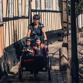 Kvinde kommer kørende på ladcykel med to børn i på en smal, brostensbelagt gade med Bybækken til den ene side og gamle bindingsværkshuse til den anden side.