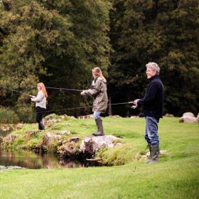 En mand, en kvinde og to børn står på en græsbeklædt flade og fisker. Der er åkander på søen og store, grønne træer i baggrunden.