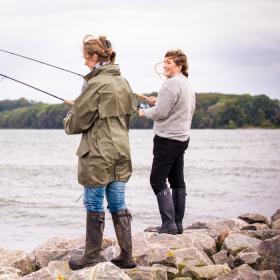 To kvinder står på store sten i vandkanten med deres fiskestang og fisker. De er iført gummistøvler , og den ene har en lang, grøn jakke på, emns den anden står i en sweater. De har begge vind i håret.