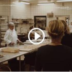 Køkkenet i OCC Odense Congress Center. To kokke står og laver mad, mens to kvinder kommer ind i køkkenet. Der er et videoikon i midten af billedet.