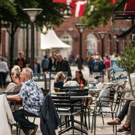 Mennesker sidder udenfor foran Lagkagehuset i Odense gågade. I venstre side står der er et skilt med med et billede af en kop varm kaffe. 