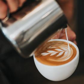 To slørende hænder er ved at lave en kop barista kaffe. I den ene hånd er en hvid kaffe kop fyldt til kanten med kaffe. I den anden hånd er en sølvkande. Fra kanden hældes der mælk ned i koppen som former et blad.