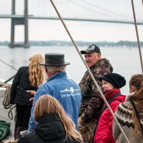 Et billede taget mellem rebbene på et sejlskib. 5 turister lytter med mens en guide med høj hat snakker. I baggrunden ses havet og Lillebæltsbroen.