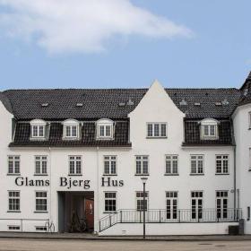 Stor, hvid bygning med gråt tegltag og tårn-lignende tilbygning med vejrhane på toppen af tårnets tag. Over døren står: Glams Bjerg Hus.