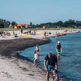 En lang strandbred. Solen skinner og midt på stranden står et rødt og hvidt livredderhus med et gult flag på toppen. På stranden er en masse mennesker i badetøj. Nogle ligger og soler på stranden, andre står i vandkanten og andre bader i stranden.