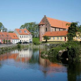 Hyggelig købstadsby med farverige huse og orange tage spejler sig i en sø. 