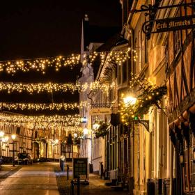 Et billede af en brostensgade i Bogense. På hver side af gaden er der forskellig farvede bindingsværkshuse. Mellem husene hænger julelys og lyser gaden op. 