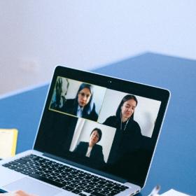 En kvinde med mørkt hår sidder med siden til og kigger ned på sin computer. På computerskærmen ses tre billeder af kvinder i et videoopkald. Computeren står på et blåt bord og ved siden af ligger en notesbog.
