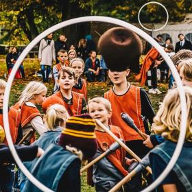 Gennem en hvid hullahopring kan man se en flok børn. Den ene halvdel har røde veste på og den anden blå. De har alle sammen kosteskafter mellem benene og kaster en bold. I baggrunden er forældre tilskuer, mens de står under et kæmpe træ. Børnene spille spillet quidditch fra Harry Potter.