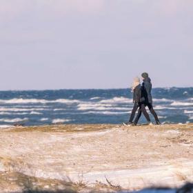 I frøperspektiv ses stranden med vandet i baggrunden og langs stranden går et par med vintertøj på