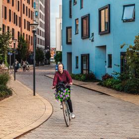 En cyklist med blomster i cykelkurven kommer cyklende i det nye kvarter 'Carl Nielsen kvarter' i Odense. Lejlighederne omkring er beige, blå, orange og bordeaux. Her ses også en masse grønne planter..  