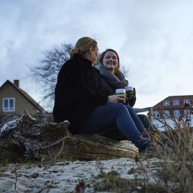 To piger sidder trærester på jorden og drikker kaffe og bag dem er bygninger i baggrunden