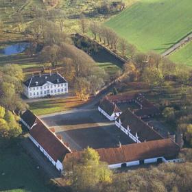 Dronebillede af Hagenskov gods. En hesteskosbygning vender mod selve godset. Bygningerne er omgivet af skov og marker. 