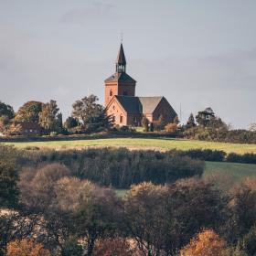 Bølgende landskab med krat, træer og grønne marker. I baggrunden troner en rød kirke frem.