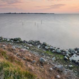 Et billede af hav med en himmel i horisonten der er oplyst i en solnedgangs farver. I forgrunden ses kyst med sten der adskiller græs fra havet.