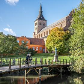 En idyllisk grøn park med en stor kirke med et spidst tårn i baggrunden. Kirken, der har en klassisk arkitektonisk stil, er omgivet af frodige træer i fuldt løv. Foran kirken er der en lille bro over en rolig å, hvor en person krydser på cykel. Himlen er klar og blå.