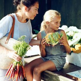 Kvinde og pige ved en åben vejbod med grøntsager. Piger holder et blomkål i hånden, kvinden et bundt gulerødder.