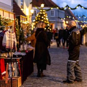 En brostensgade. Til højre er en række af små farvede huse. Til venstre er en julemarkedsbod sat op. Der er flettede kurve, børne striksweatres og julelys. Foran boden står en mand og kvinde og taler. I baggrunden kan man spotte flere slørede mennesker og boder.