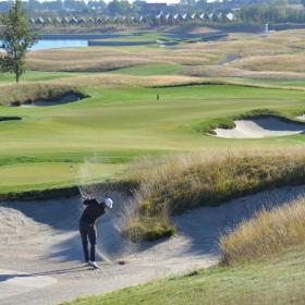 Golfspiller på bølgende grøn golfbane slår et slag fra en bunker, så sandet står op.