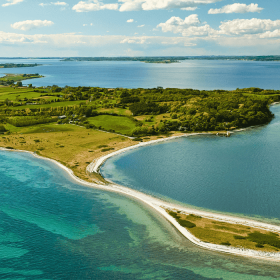 Drone photo of Avernakø in sharp, lush, green colors with oceanblue water all around the shores and the southfunen archipelago in the horizon