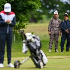 En golfspiller står med sin golftaske på en græsplæne med to tilskuere i baggrunden