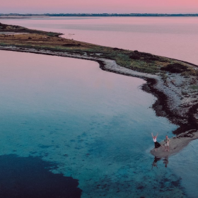 Et par står for enden af en landtange på et dronebillede med solnedgangen i baggrunden