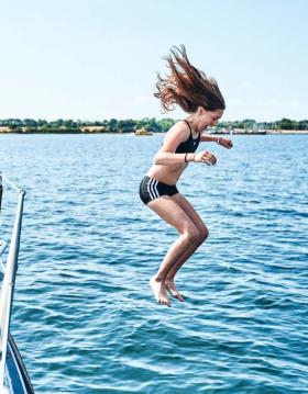 Pige midt i et hop fra båd. Det er sommer, havet er blåt og solen skinner.
