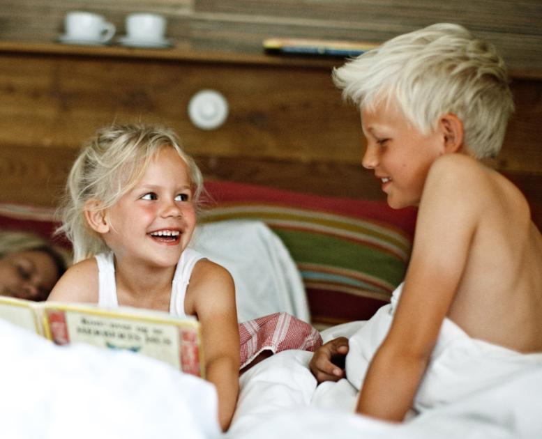 En lyshåret pige og dreng sidder i en seng, kigger på hinanden og griner. Pigen sidder med en bog i hånden.
