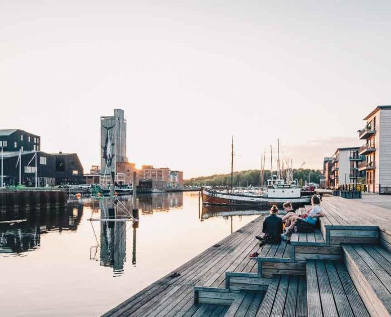 Stemningsbillede fra Odense havn en aften med solnedgang. Mennesker sidder på en træplatform ud til vandet og kigger på skibene.