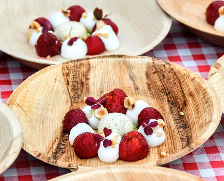 På en rød og hvid ternet dug står otte bæredygtige paptallerkner. På tallerknerne er en flot anrettet dessert bestående af jordbær, hvidt skum og en iskugle i midten.
