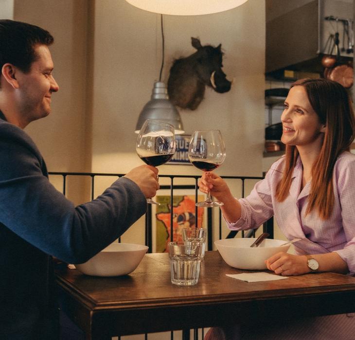 En kvinde og en mand spiser på en restaurant. Der står tallerkener på deres bord og de smiler og skåler i rødvin.