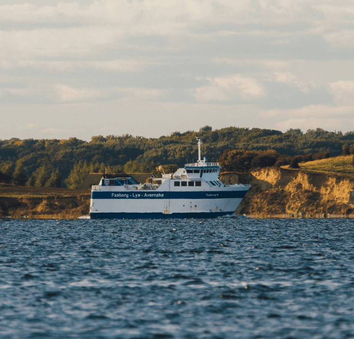 En hvid og blå færge sejler over et farvand i det maleriske Sydfynske øhav med en kuperet, grøn kystlinje i baggrunden under en overskyet himmel.